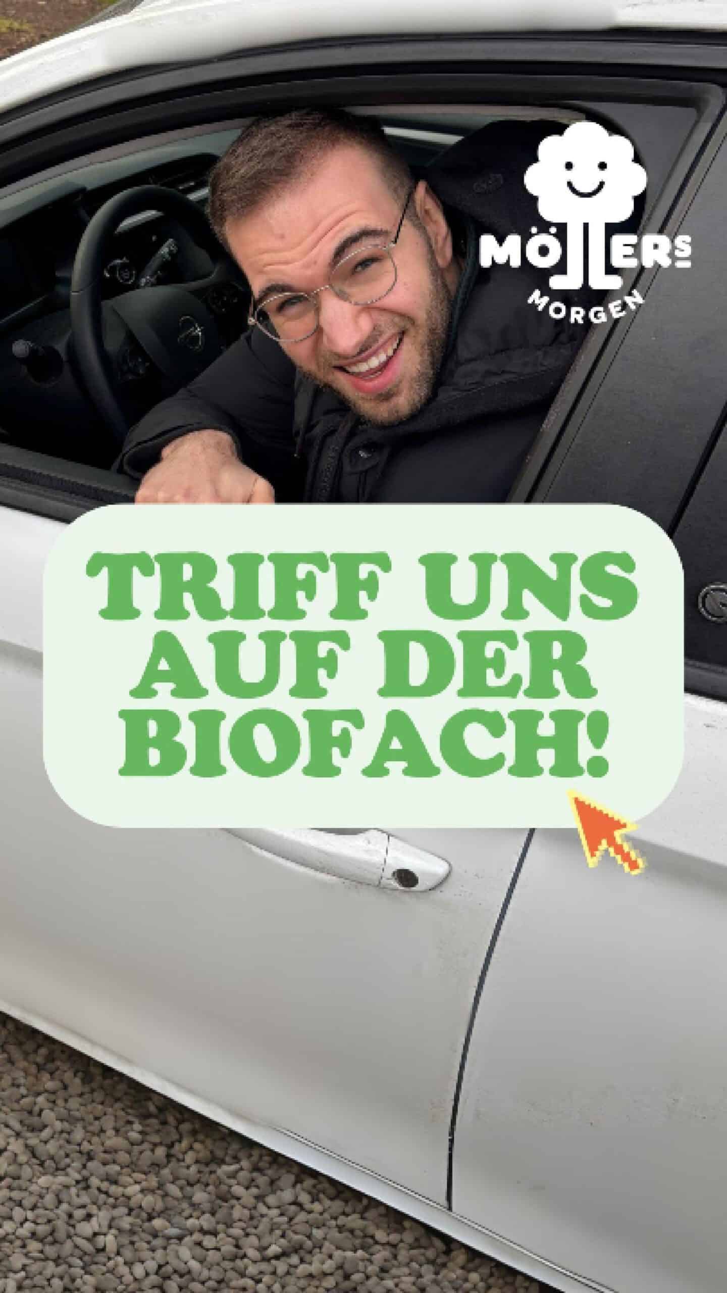 Person in einem schwarzen Wintermantel schaut aus einem weißen Auto heraus und lächelt. Der Text „TRIFF UNS AUF DER BIOFACH!“ in grünen Buchstaben ist über dem Bild zu sehen, zusammen mit einem Cursor-Pfeil. Das Möllers Morgen-Logo befindet sich oben links.